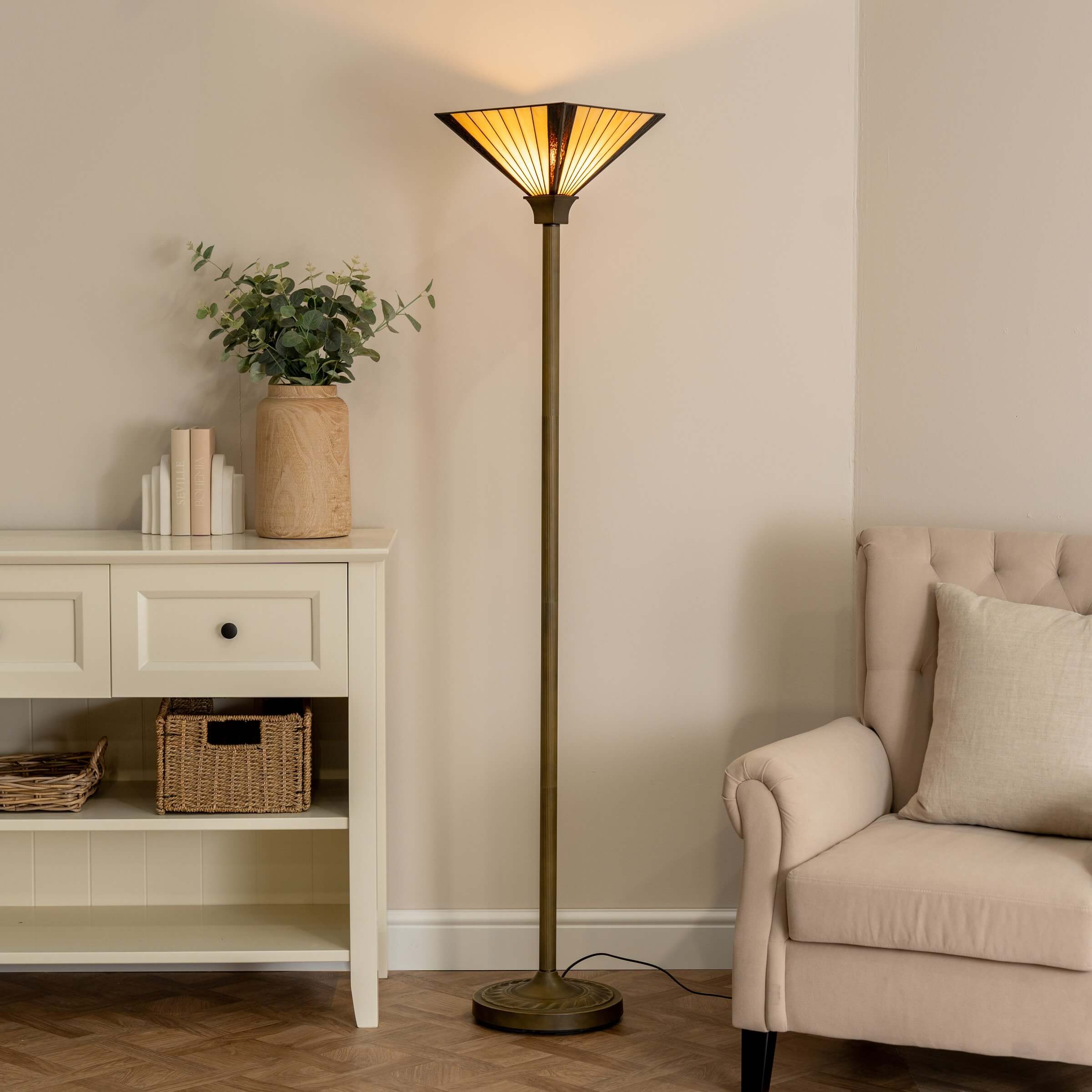 Amber-shaded floor lamp with antique brass stem stands beside a cream console and tufted armchair in a softly lit, neutral-toned living room.