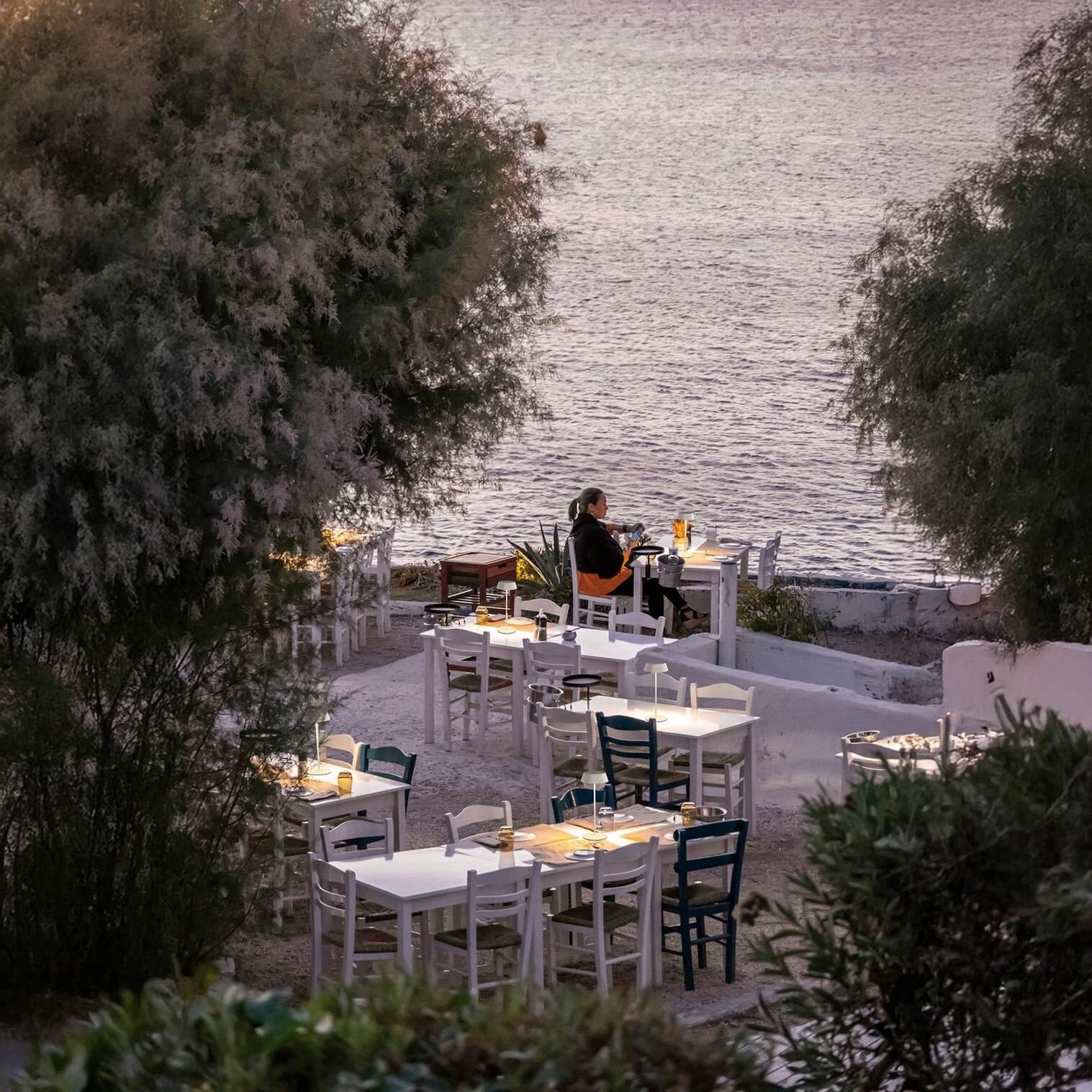 Small glowing table lamps illuminating seaside dining tables at dusk.