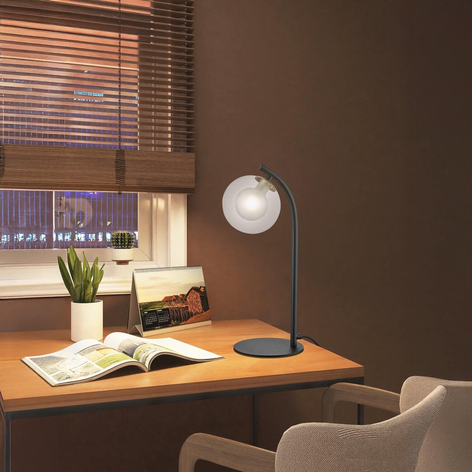 Cozy workspace with a wooden desk, modern lamp, open magazine, and potted plant by a window with blinds.