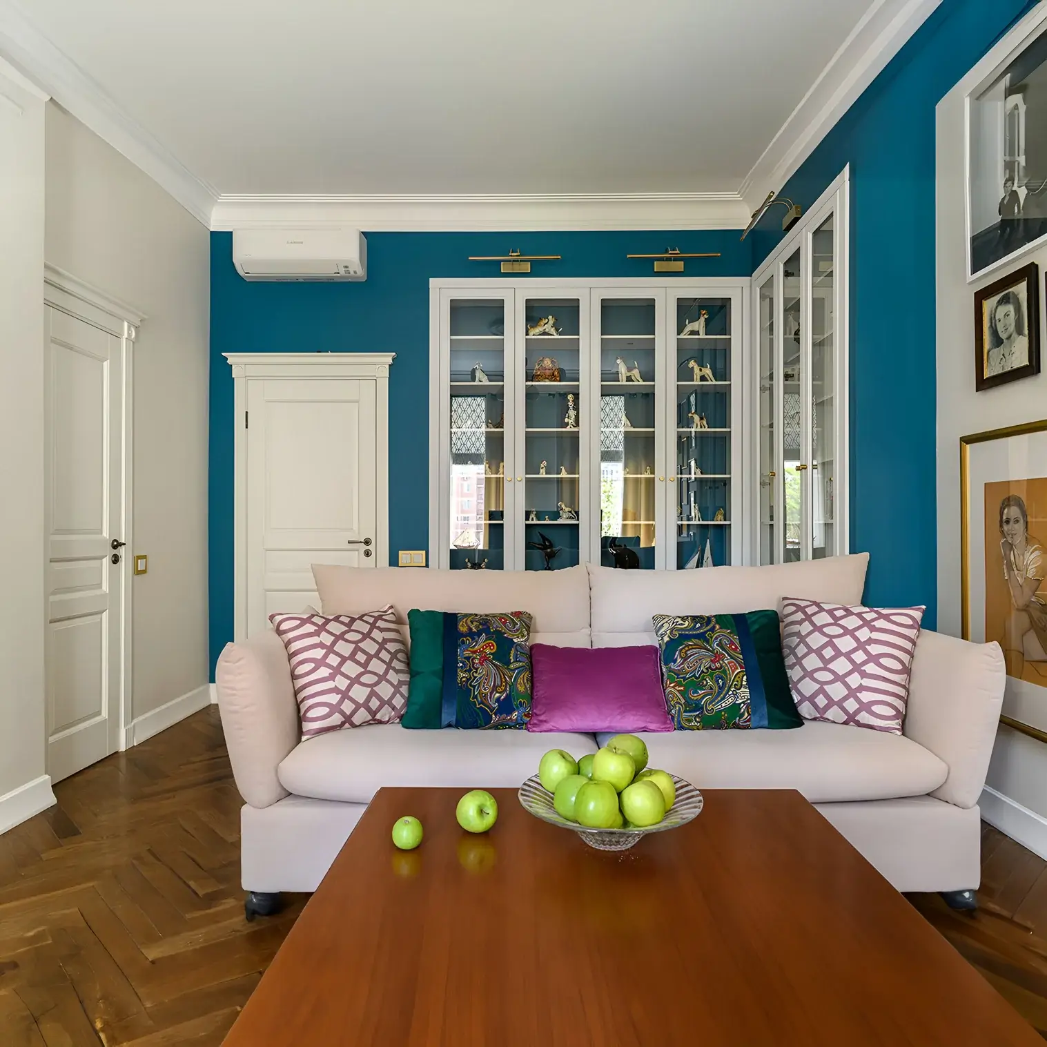 A vibrant living room featuring a bold blue accent wall, glass-front cabinets with collectibles, and a cream sofa adorned with colourful patterned cushions. Picture lights highlight the display shelves.