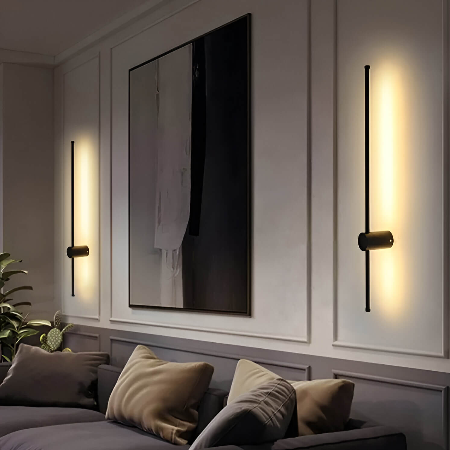 A cozy living room with vertical wall-mounted lights casting soft ambient lighting. The lights frame a large black and white artwork above a grey sofa with neutral-toned cushions.