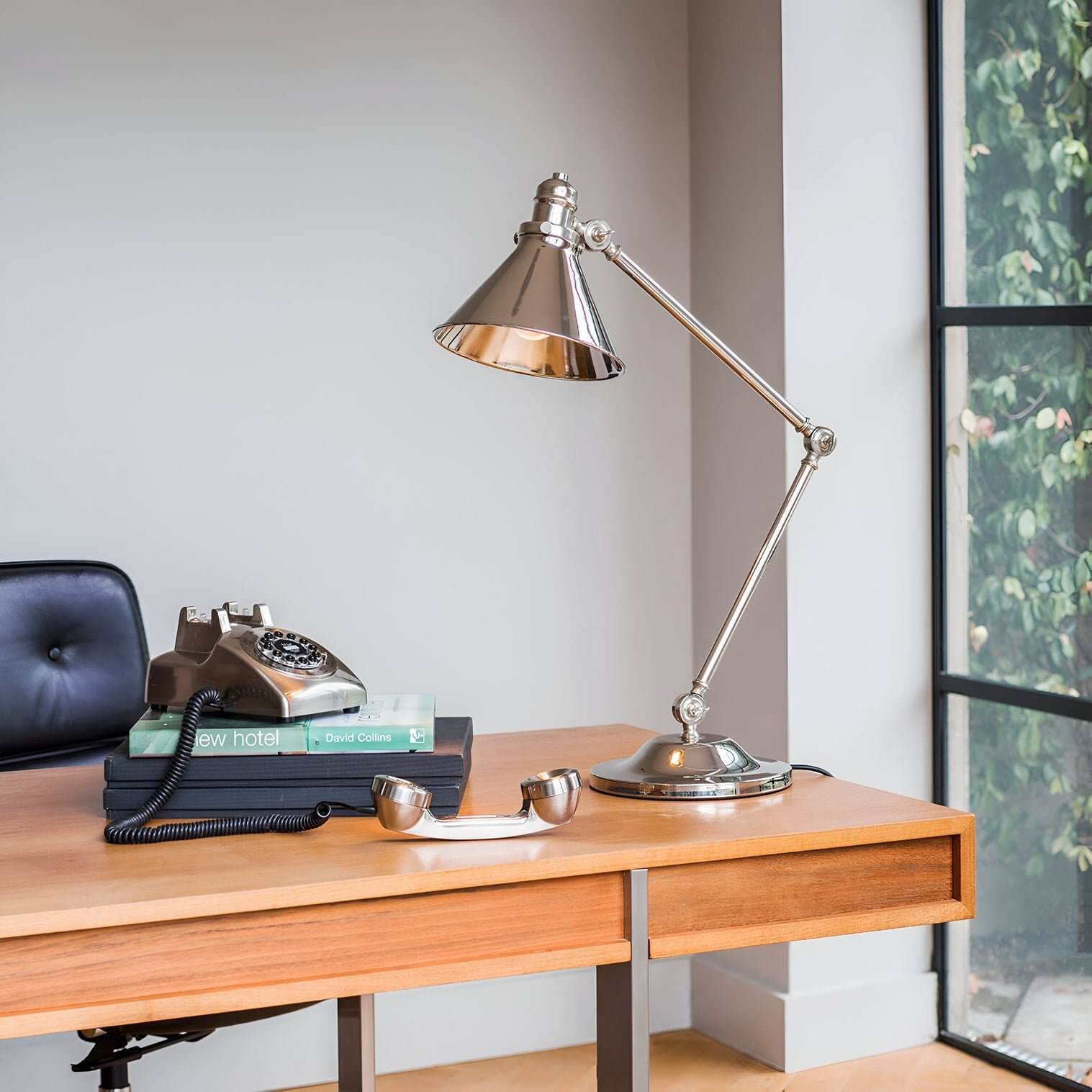 Adjustable polished metal desk lamp with conical shade on a wooden desk beside a vintage telephone.