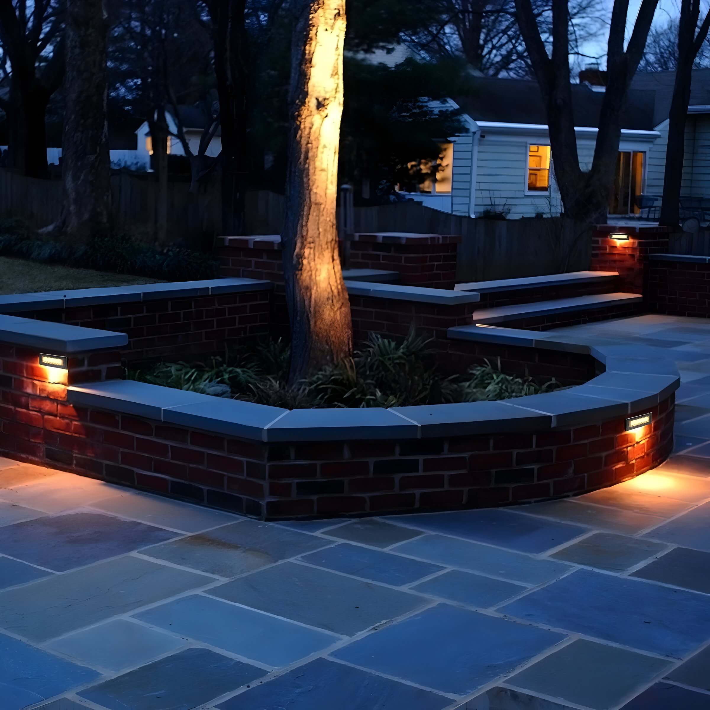 Curved red brick seating walls with rectangular wall lights illuminating a tree-centered patio at night.