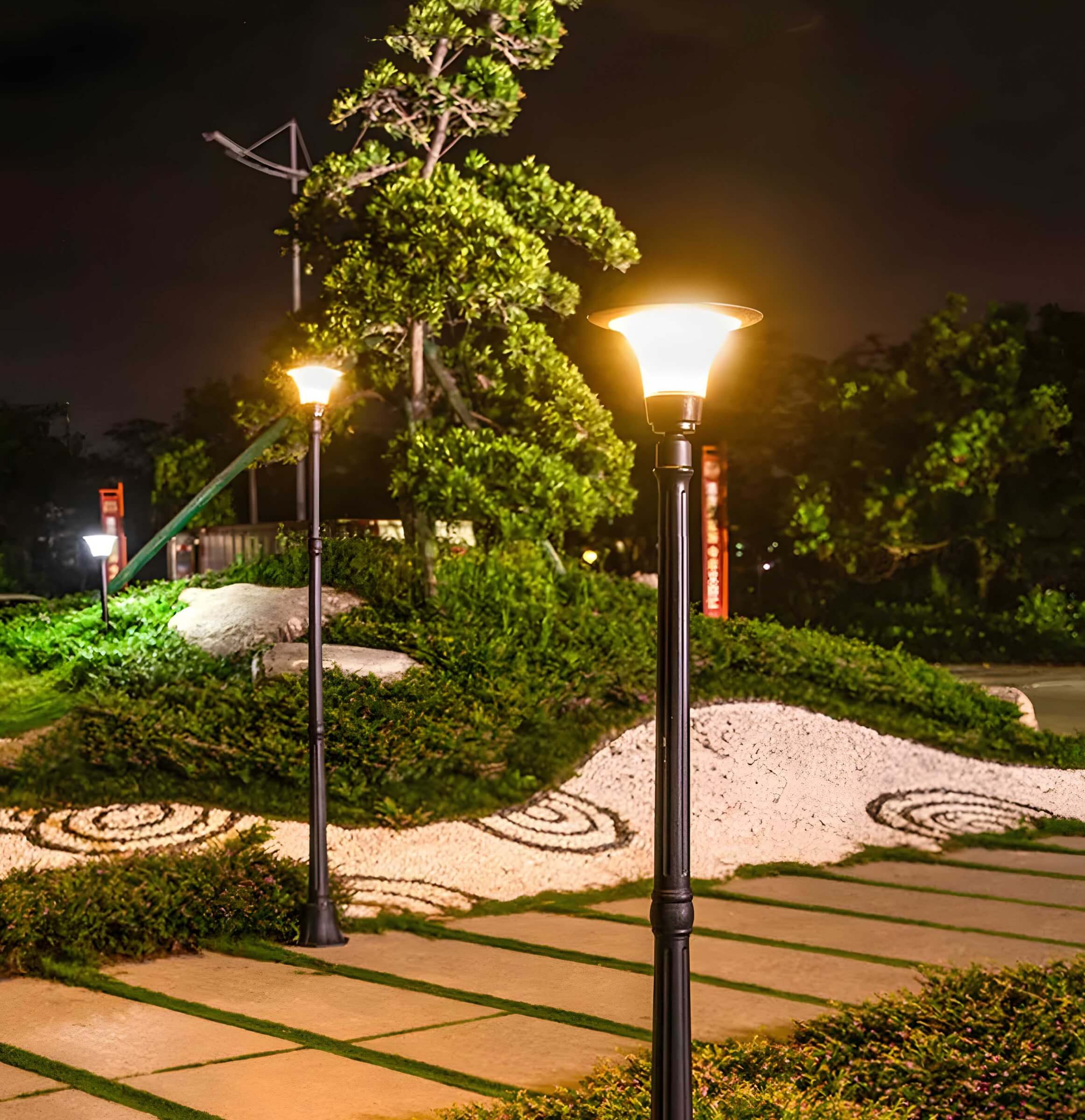 Illuminated black lamp posts at night along a landscaped walkway with sculpted greenery.