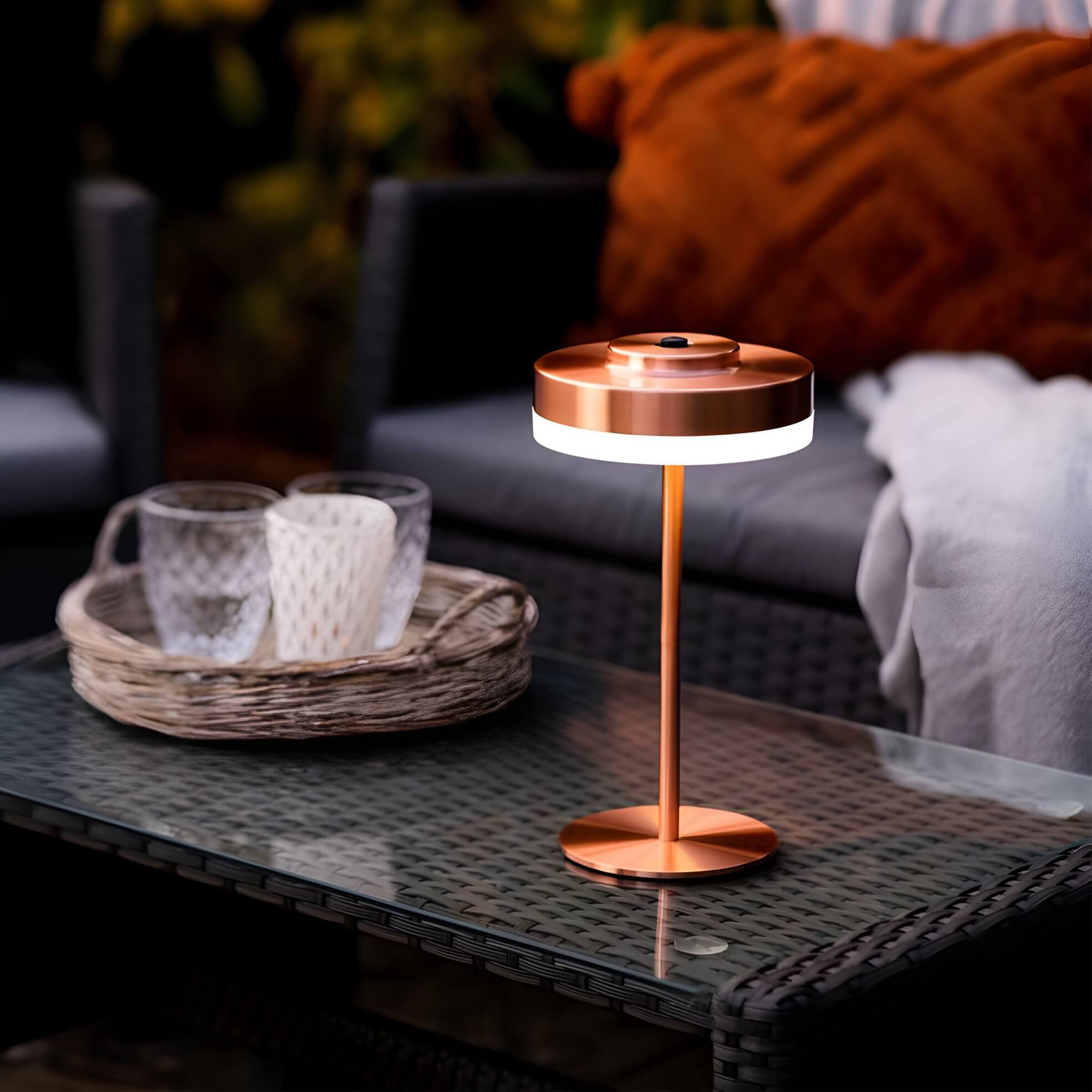 Copper-toned portable LED table lamp on a glass-top wicker coffee table with glassware and a rust cushion in the background.