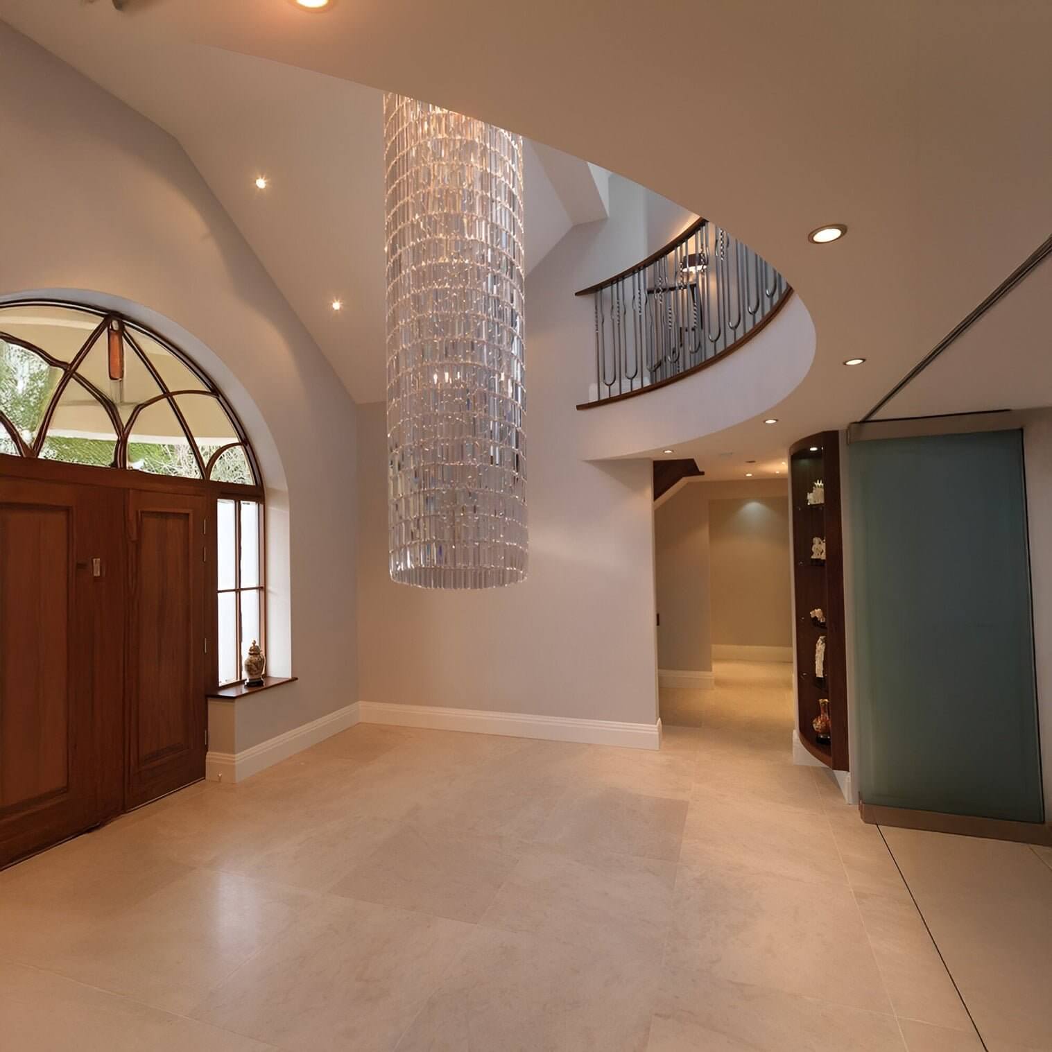 Tall cascading crystal chandelier in a cylindrical design hangs in a grand double-height entryway with curved staircase.