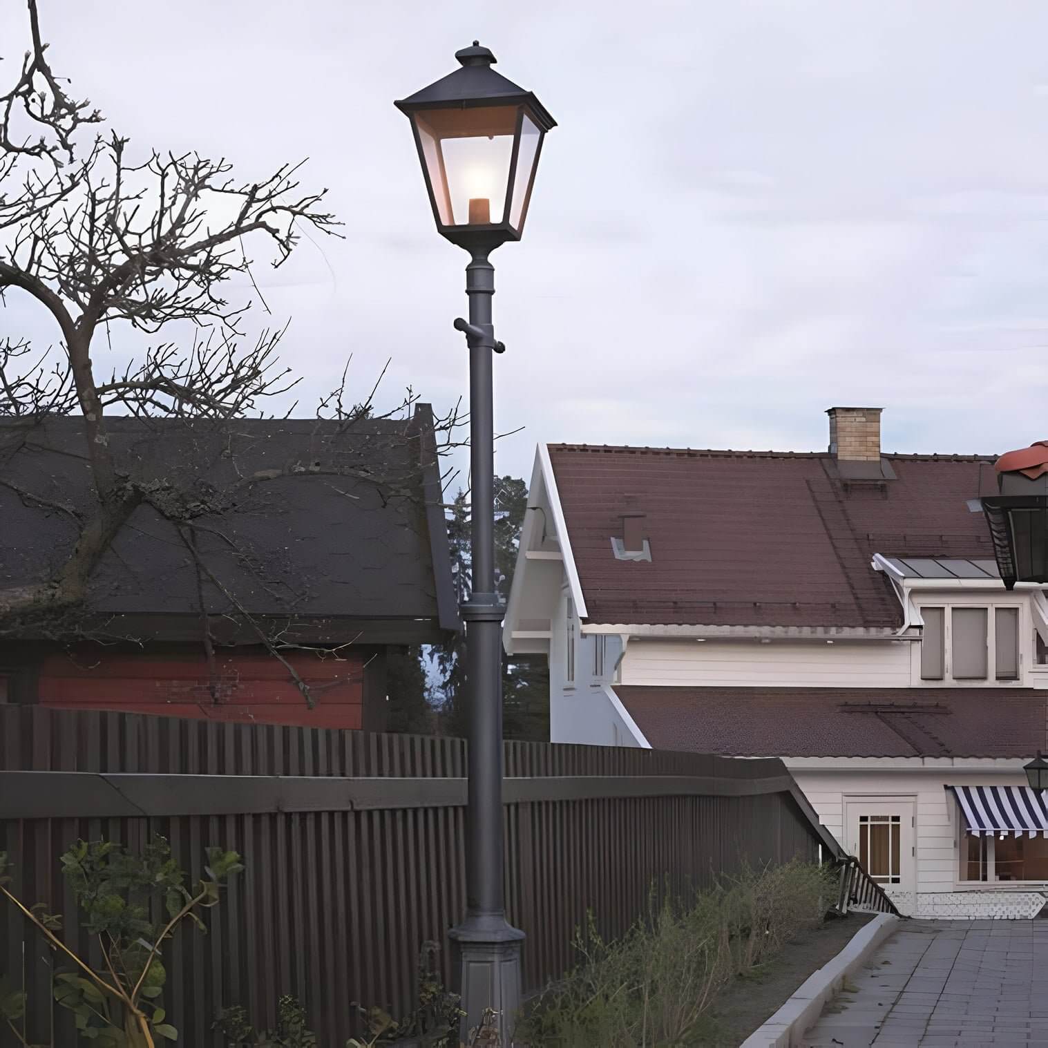Classic black lamp post with lantern head along a paved sidewalk near white house and sloped fence at dusk.