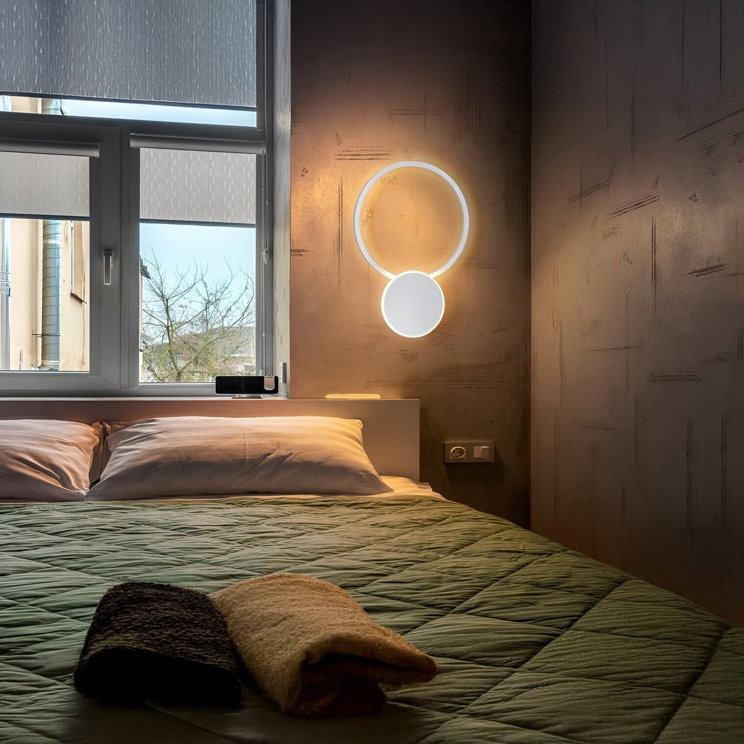 Minimalist circular LED wall fixture with halo effect, mounted beside a bed with green quilted bedding and folded towels.