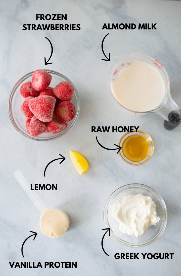A photo of ingredients for strawberry cheesecake protein smoothie laying flat against a white marble surface.