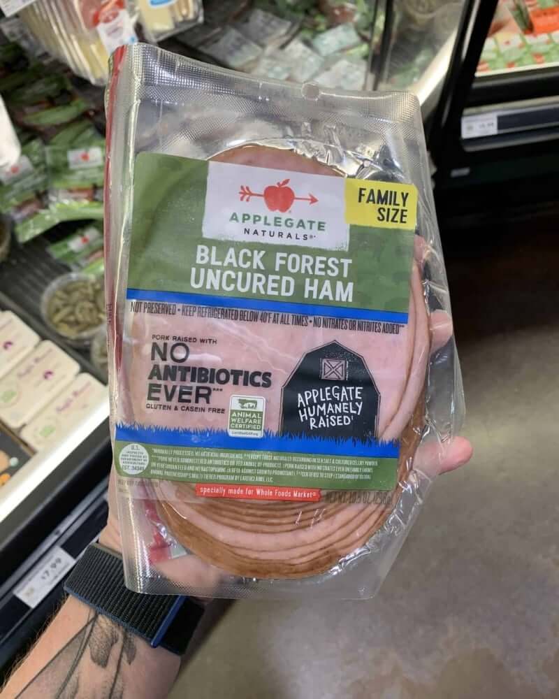 A hand holding a package of deli meat branded Black Forest Uncured Ham from Applegate.