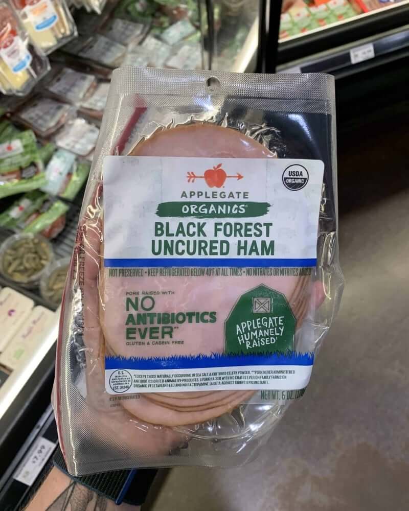 A hand holding a package of deli meat branded Applegate | Organics Black Forest Uncured Ham. 