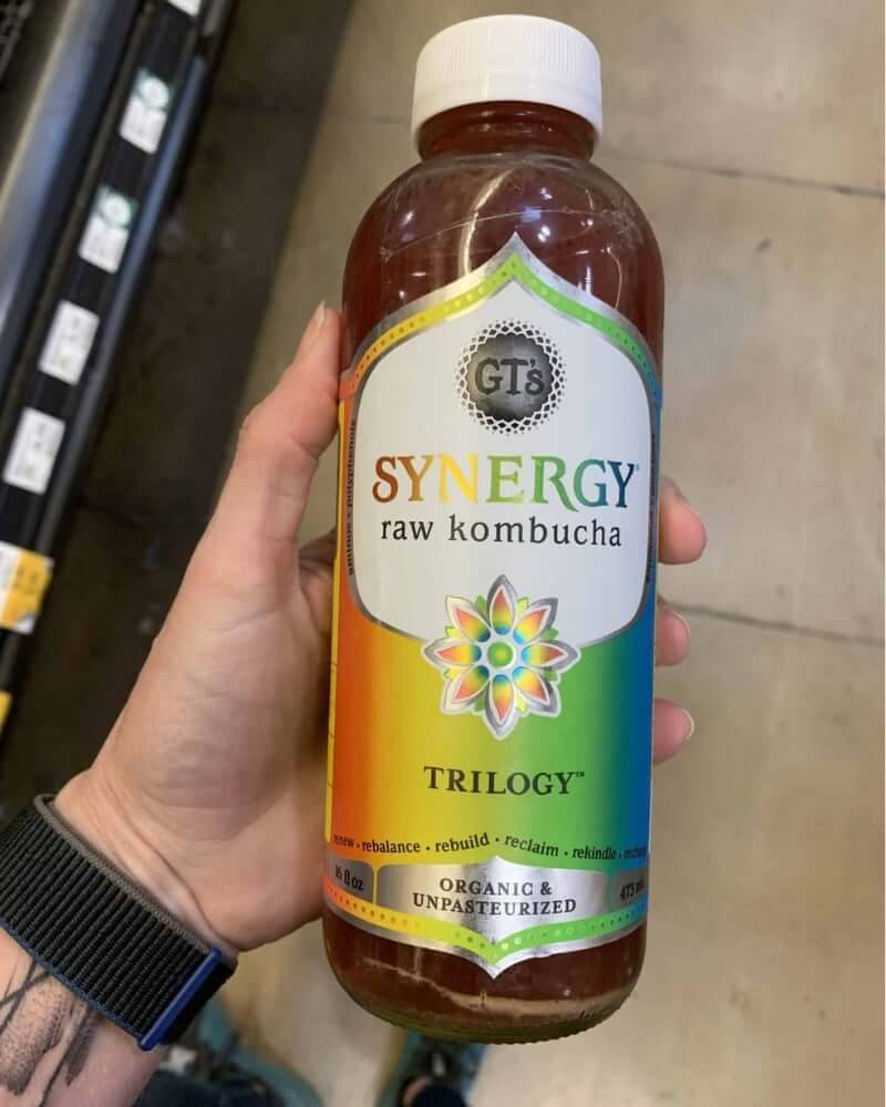 A photo of a hand holding a kombucha bottle labeled Synergy Raw Kombucha.