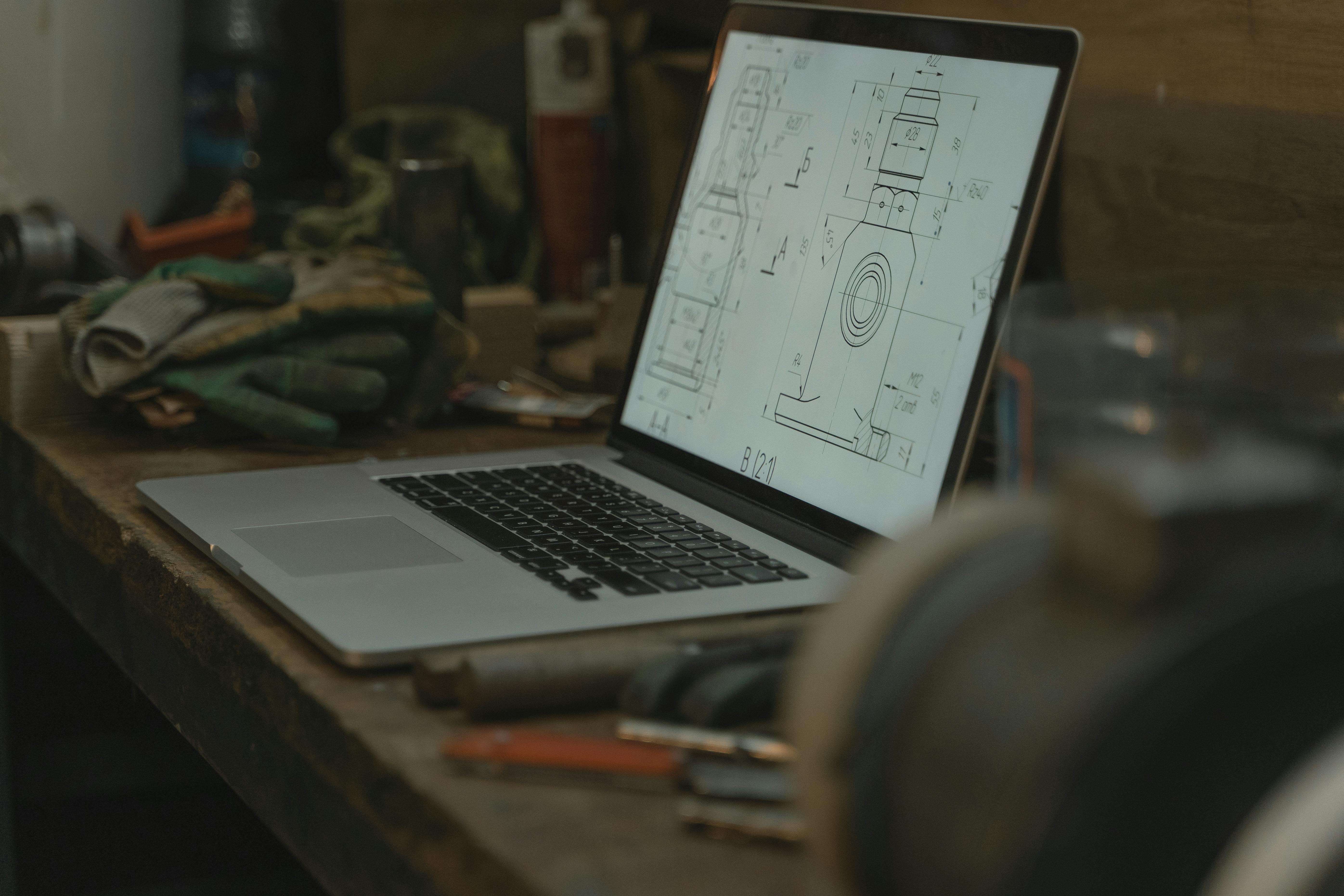 Laptop showing a technical drawing, surrounded by tools and gloves in a workshop setting.