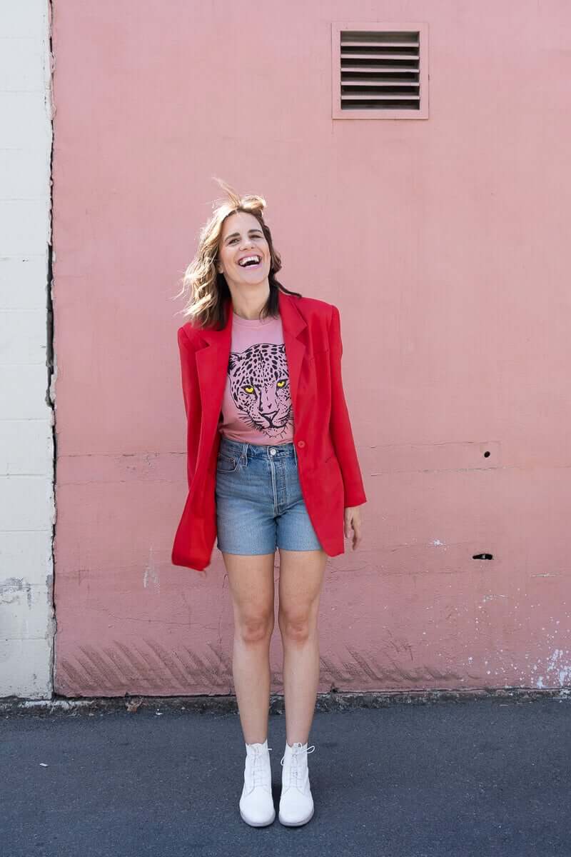 Angela wearing the Thread Hippie Pink Leopard women’s graphic tee styled with a bright red blazer and denim cut-off shorts.