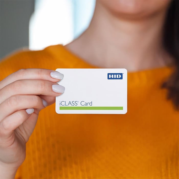 Alt text: A woman wearing a mustard yellow sweater holds an HID iCLASS access card between her fingers, displaying the front of the card with the HID logo and green stripe.