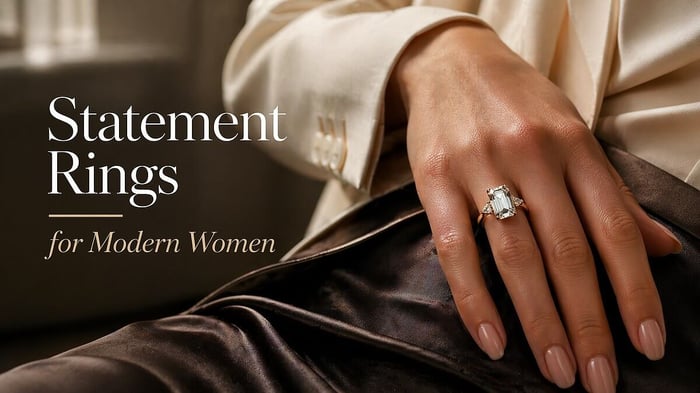 Statement rings: Close-up of a modern woman’s hand resting lightly on a tailored cream blazer and dark silk skirt, wearing one striking natural diamond statement ring with an elongated centre stone and refined gold setting.