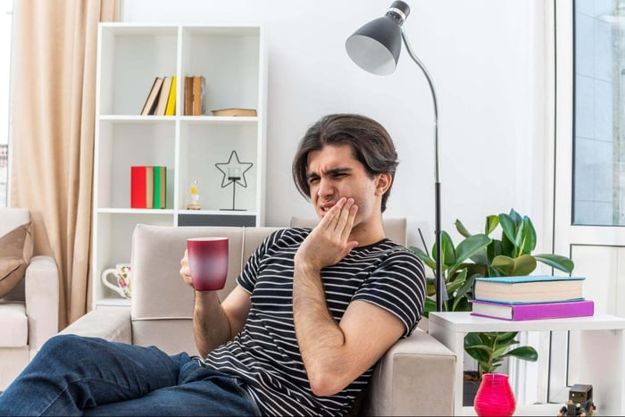 Young man holding jaw in pain while sitting with a coffee mug.