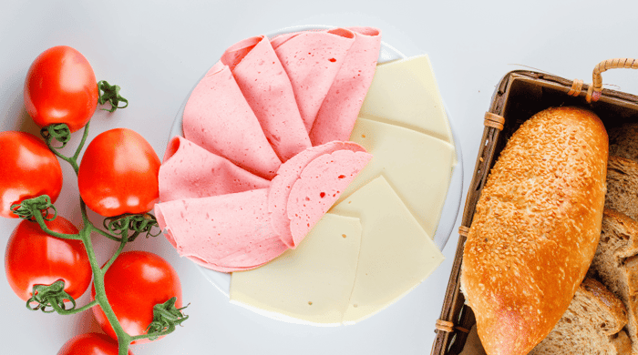 High protein food spread with sliced bologna and white cheese fanned on a plate served alongside vine tomatoes and a sesame seed baguette.