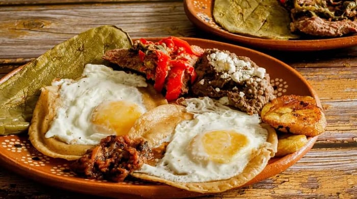 High protein meals featuring a traditional Mexican breakfast plate with two fried eggs, refried beans, grilled meat, roasted peppers, fried plantains, and tortillas on a clay plate.