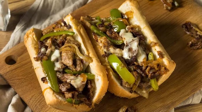 High protein Philly cheesesteak with two open hoagie rolls loaded with shaved steak, sautéed green peppers, caramelized onions, and melted white cheese on a wooden board.