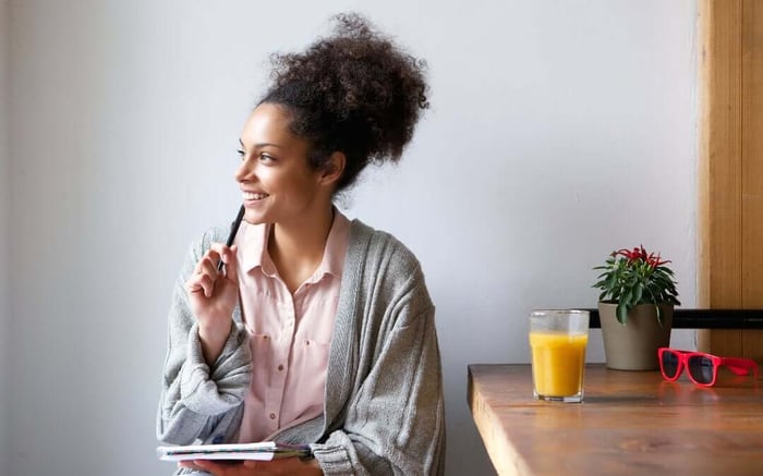 woman sitting at desk practicing decision making confidence with notepad, holding pen, plant, orange juice, red sunglasses