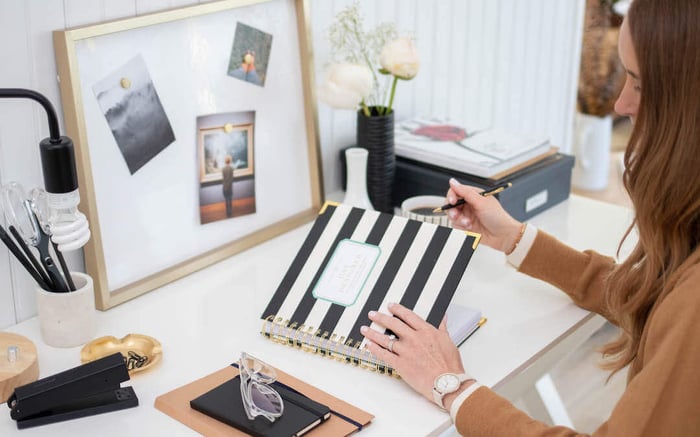 woman holding black and white striped planner, learning how to prioritize your life, desk, whiteboard with pictures, lamp, office supplies, vase with flowers