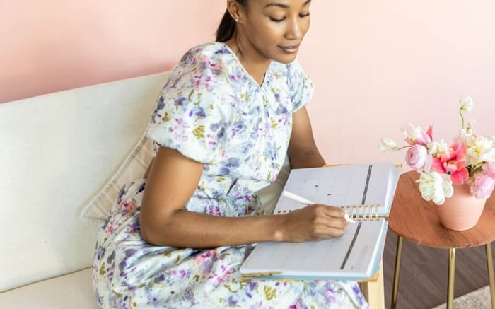 woman sitting wearing a floral dress, holding an open book, getting organized with your new planner, table with vase and flowers
