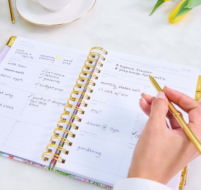 planning your academic year, hand holding gold pen, open book planner showing written on pages, coffee cup, yellow flowers, white background