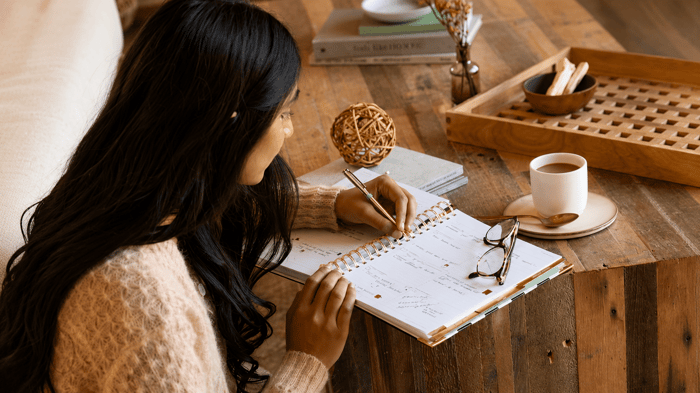 Planning Ahead for a Happier, More Intentional Life, woman writing in planner, glasses, coffee cup, wood table, tray, books, vase