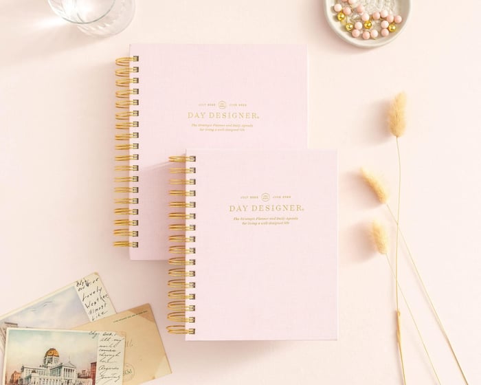 Which Planner Is Right For You?, stacked blush pink planners to show sizing options, dried wheat, dish with beads, postcards, water glass, beige background