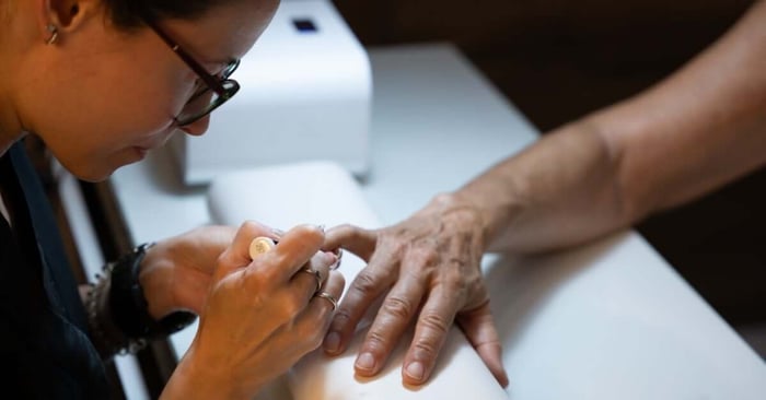 A female nail technician is holding a brush and applying nail polish to a client while sitting at a white table.