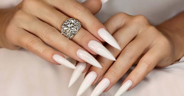 A close-up of a woman's stiletto nails. Her nails are white and extra long. She's also wearing a cluster diamond ring.