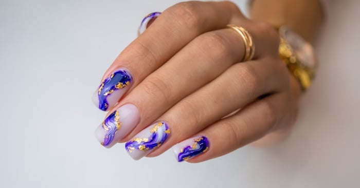 A close-up on a person's nail art. Their nails have a white base with painterly blue streaks and gold flaking.