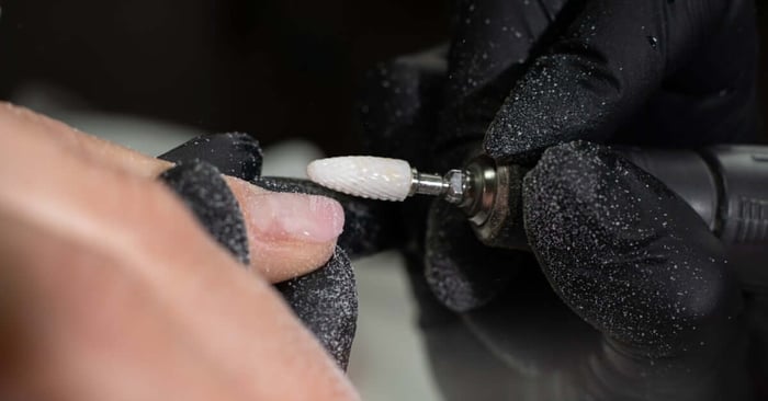 A professional wearing black gloves and using a tool bit to grind away the surface of a client's nail.