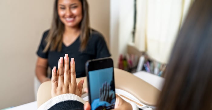 A client taking a photograph of their manicured nails while a smiling technician sits in the background.