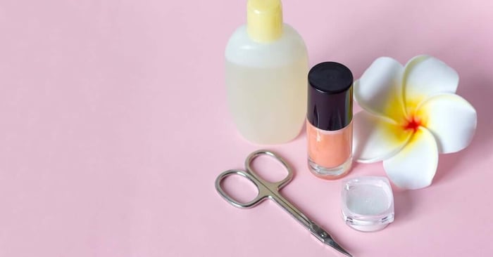 Nail polish, nail remover, scissors used for manicures and pedicures, and a flower are all sitting on a pink background.