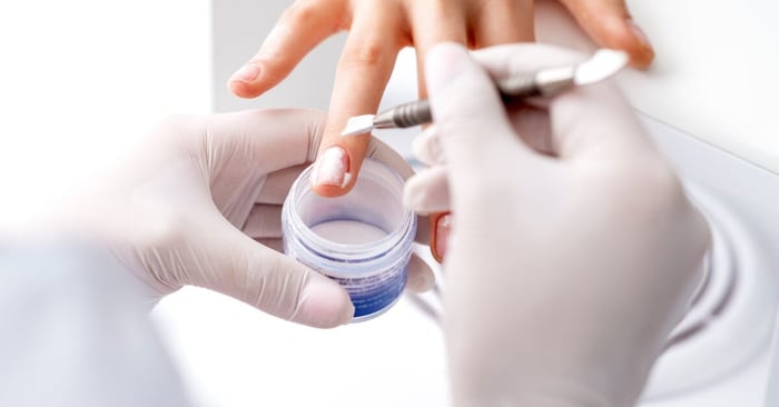 A close-up of a nail tech's gloves applying acrylic powder on the nails of a young person in a beauty salon.
