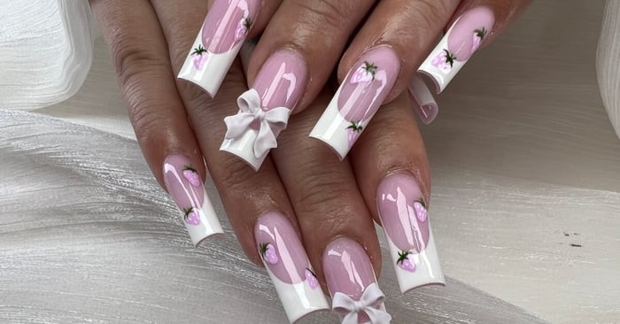 A manicured hand displaying long 3D acrylic nails with pink polish, white tips, floral designs, and dimensional bow accents.