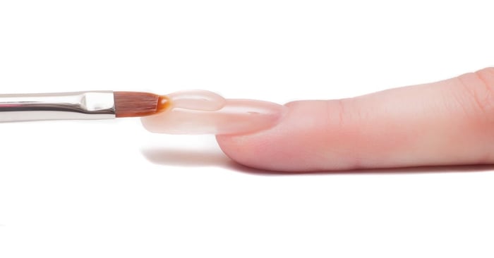A close-up view of a manicurist applying a bead of acrylic gel to the top of a light-pink nail using a clean brush.