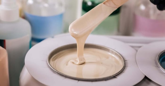 A wax spatula drips warm cream wax into a heated wax warmer, with salon products blurred in the background.
