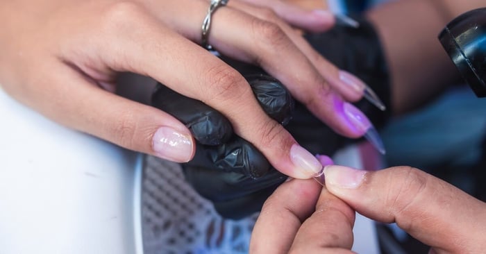 A manicurist working in a salon environment wears black gloves and applies a soft-gel tip to a nail surface using adhesive.