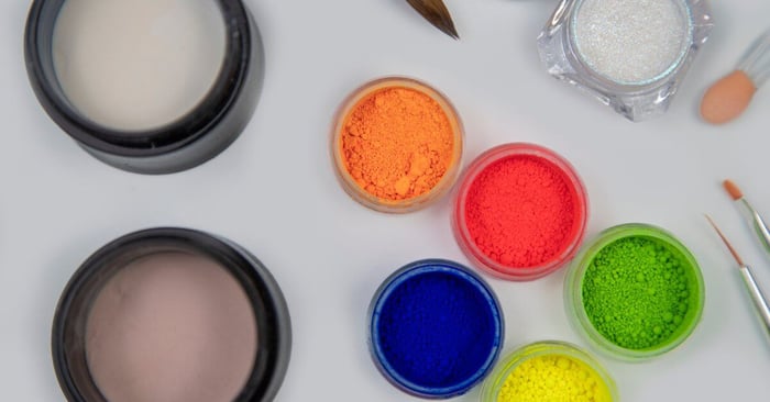 Nail tools rest on a table alongside orange, green, red, blue, yellow, tan, and white acrylic powder.