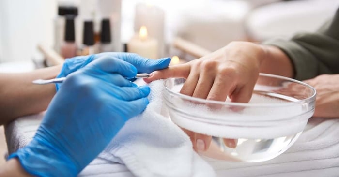A manicurist uses a cuticle pusher while a young woman soaks her hands in nail bath at a professional salon.