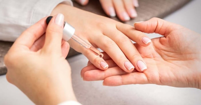 A close-up of a manicurist applying oil to a person's cuticles using a pipette. The person has a French manicure.