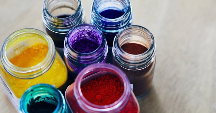 A close-up of opened jars of various sizes filled with colored powders spread out on a light-colored table.