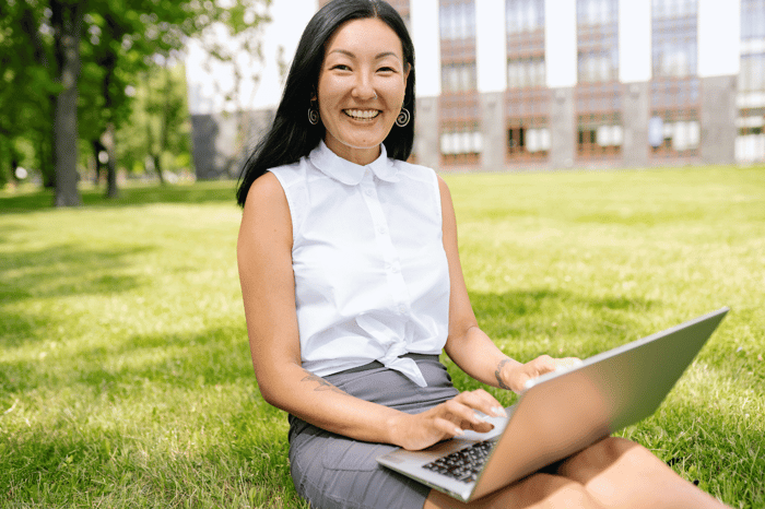 Smiling transfer student sitting on campus lawn using a laptop while researching how to choose the right online college.