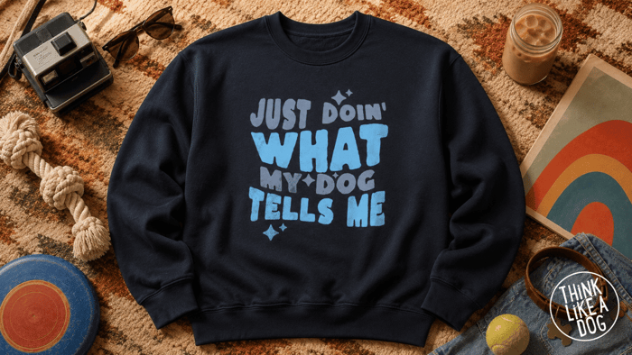 Black crewneck sweatshirt with blue “Just Doin’ What My Dog Tells Me” graphic, styled flat lay with dog toys, iced coffee, denim, and retro props.