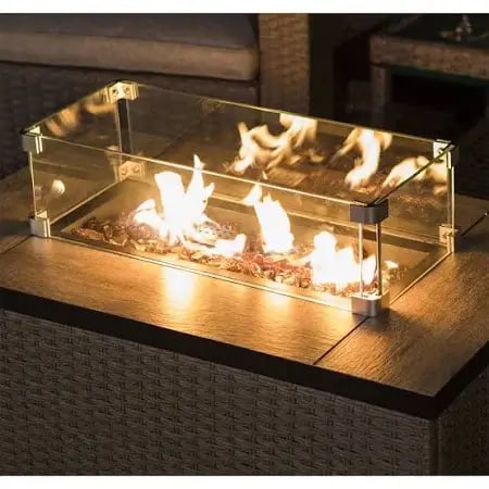 A rectangular fire pit table with a glass wind guard. The fire pit is filled with fire wood and flames are visible through the glass wind guard. The table is surrounded by a wicker seating area.