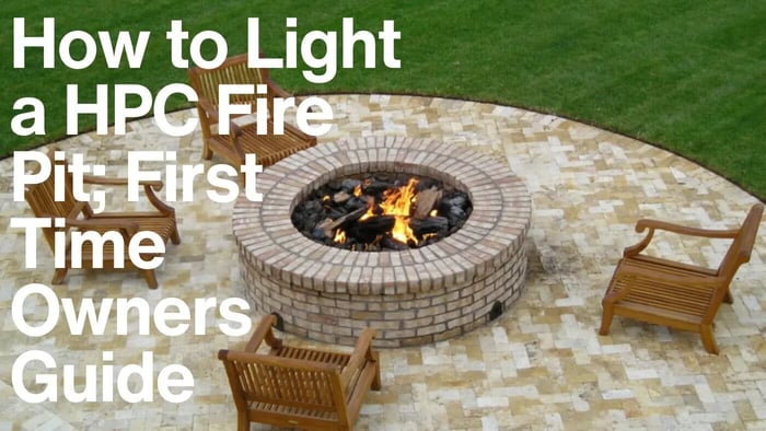 A wide shot of a modern outdoor living area featuring a round concrete fire pit with a flame, surrounded by a curved concrete bench. In the background, there are three dark pergolas and trees.