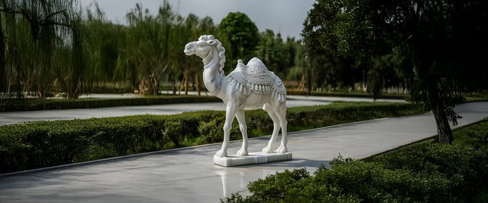 White camel statue on a paved walkway, part of an outdoor sculpture display in a lush green park.